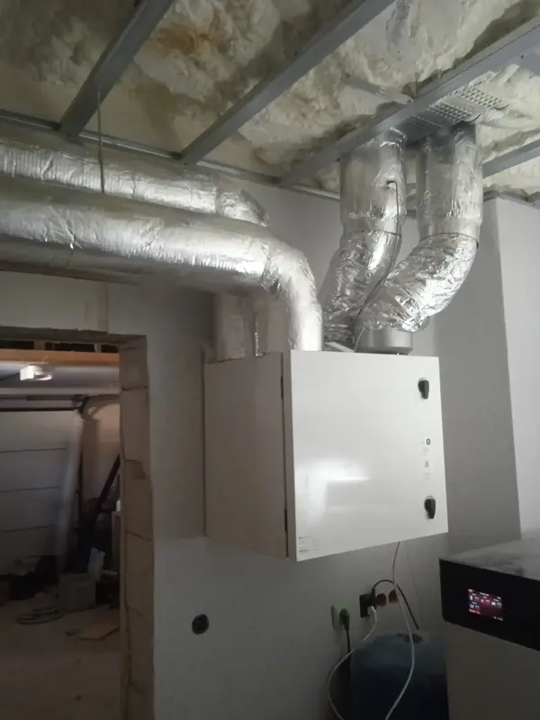 Rekuperator wiszący na ścianie, od którego odchodzą grube, aluminiowe rury