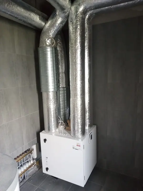 Rekuperator zamontowany w kącie pomieszczenia. Z rekuperatora odchodzą srebrne, grube rury